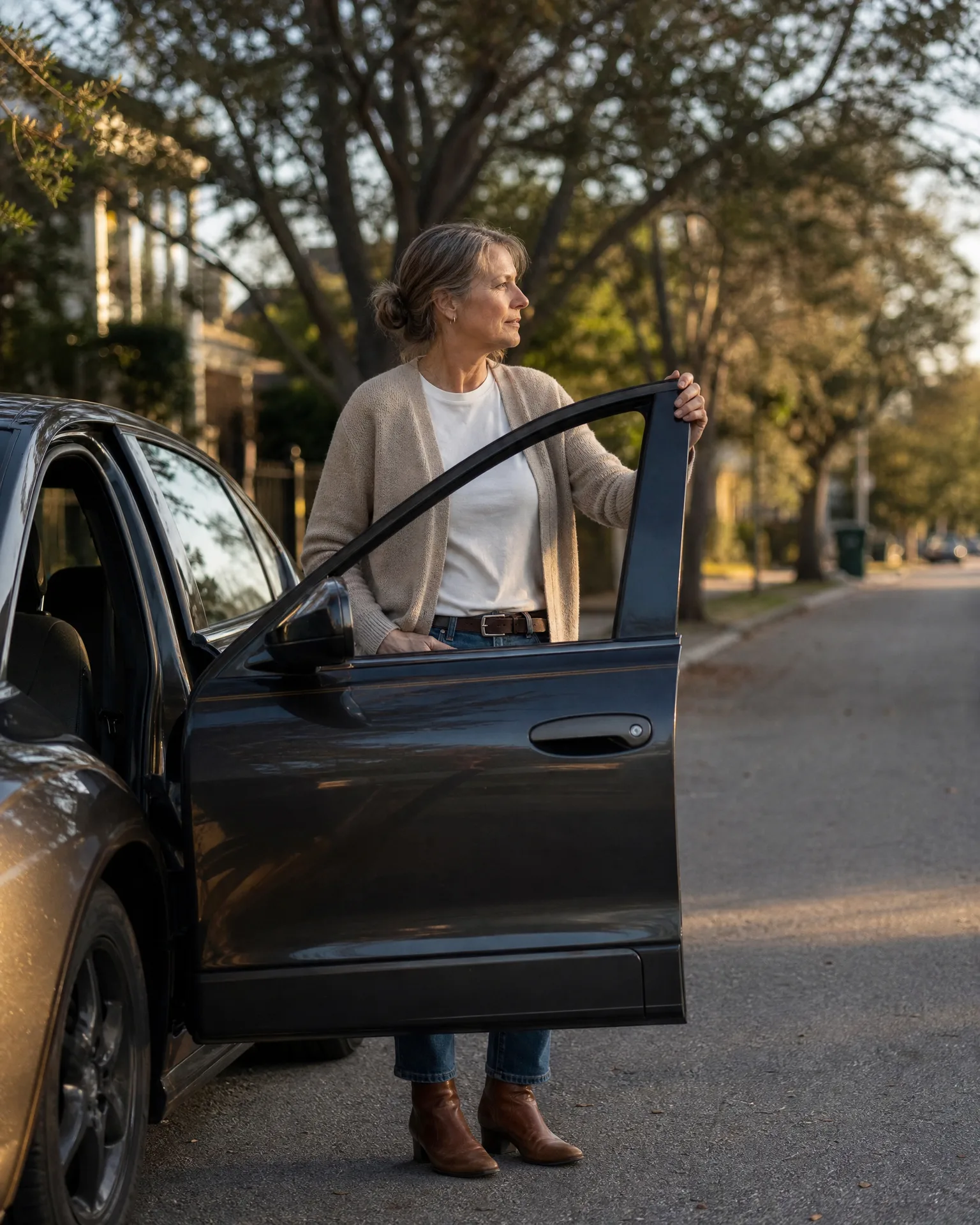 A woman in a beige cardigan, white top, jeans and brown ankle boots stands beside a dark sedan with the open front door, looking off to the side on a tree-lined residential street.
